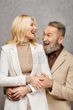 Mature man and woman, elegantly dressed, pose next to each other in a loving manner on a gray backdrop.
