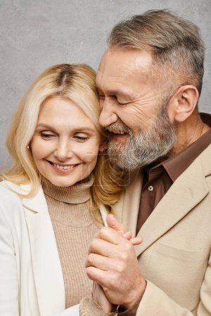 Un homme et une femme mûrs, vêtus élégamment, partagent un câlin tendre sur un fond gris.