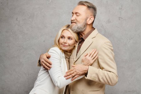A mature loving couple in debonair attire warmly embrace each other in a graceful pose against a gray backdrop.