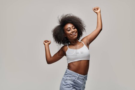 Young African American woman with curly hair dancing gracefully in a white top in a studio setting.