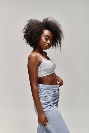 Photo for A beautiful young African American woman with curly hair poses in a white top and blue jeans in a studio setting. - Royalty Free Image