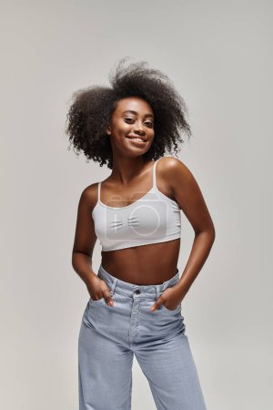 Photo for A stunning African American woman with curly hair wearing a white crop top and jeans poses in a studio setting. - Royalty Free Image