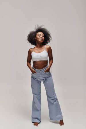 Photo for A beautiful young African American woman with curly hair elegantly poses in a white top and blue pants. - Royalty Free Image