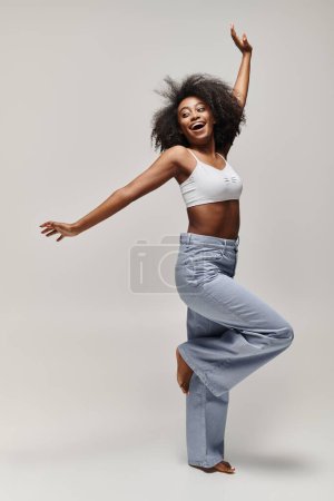 Eine schöne junge Afroamerikanerin mit lockigem Haar tanzt energisch in einem weißen Top in einem Studio-Setting.