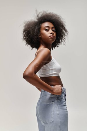 Photo for A beautiful young African American woman with curly hair poses confidently in a white top and jeans. - Royalty Free Image