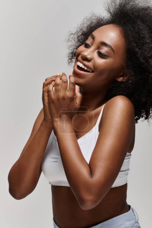 Eine schöne junge Afroamerikanerin mit lockigem Haar trägt ein weißes Oberteil, lächelt strahlend und klatscht in die Hände.