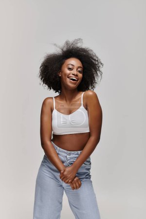 Photo for A stunning young African American woman with curly hair, wearing a white top and blue jeans, exudes elegance in a studio setting. - Royalty Free Image