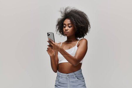 Eine atemberaubende Afroamerikanerin mit lockigem Haar hält ein Handy in der Hand, während sie in einem Studio ein weißes Crop Top trägt.