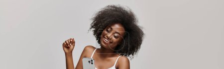 A beautiful young African American woman with curly hair holding a cell phone while wearing a white tank top.