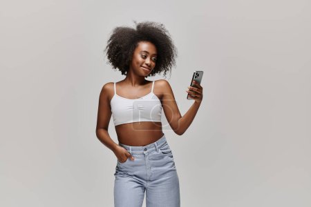 Téléchargez les photos : Une femme afro-américaine élégante avec des cheveux bouclés portant un haut de culture, tenant un téléphone portable dans une pose à la mode. - en image libre de droit