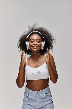 Photo for A beautiful African American woman with curly hair stands before a gray backdrop, wearing headphones. - Royalty Free Image