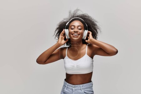 Téléchargez les photos : Une jeune afro-américaine aux cheveux bouclés écoute attentivement à travers des écouteurs dans un haut blanc. - en image libre de droit