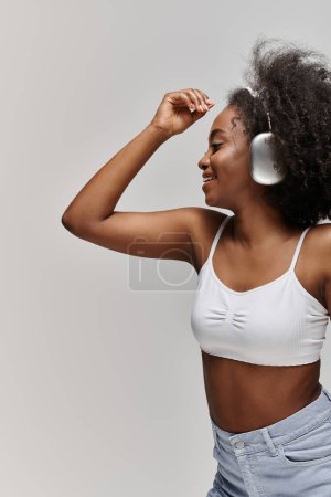Photo for A stunning African American woman with curly hair in a white bra top stylishly holds a hair dryer. - Royalty Free Image