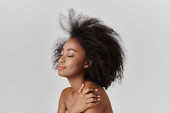 A beautiful young African American woman with curly hair stands nude as her hair cascades in the wind, exuding grace and beauty. Poster #702027066