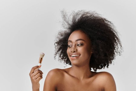 Foto de Una joven afroamericana con el pelo rizado sostiene un cepillo, centrándose en el cuidado de la piel y la belleza en un entorno de estudio. - Imagen libre de derechos