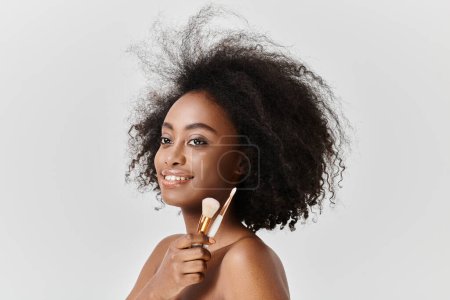 Foto de Una joven afroamericana confiada con el pelo rizado sosteniendo pinceles de maquillaje en su mano. - Imagen libre de derechos