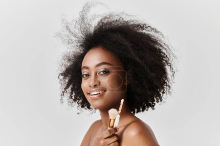 Photo for African American woman with curly hair holding makeup brushes, applying makeup in a studio setting. - Royalty Free Image