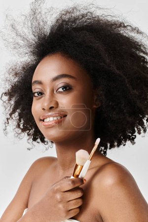 Téléchargez les photos : Une jeune afro-américaine aux cheveux bouclés tenant des pinceaux de maquillage, mettant en valeur le concept de soins de la peau dans un cadre studio. - en image libre de droit