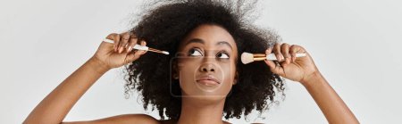 Young African American woman with curly hair styling herself using a brushes in studio setting.