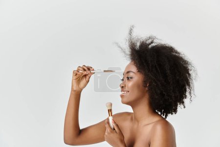 Photo for A young African American woman with curly hair applying a brush to her face as part of a skin care routine. - Royalty Free Image