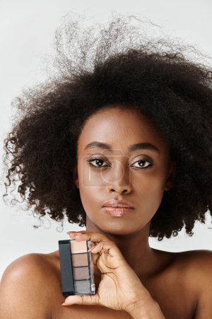Téléchargez les photos : Une jeune Afro-Américaine à la coiffure afro bouclée tient une palette de maquillage, mettant en valeur sa routine de soins de la peau. - en image libre de droit
