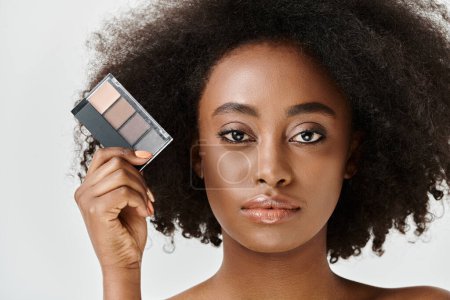 A beautiful young African American woman, with curly hair, holds a palette of makeup in front of her face.