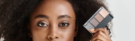 Photo for A woman with curly hair holds a makeup palette in front of her face, showcasing artistry and beauty in studio setting. - Royalty Free Image