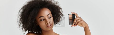 Téléchargez les photos : Une belle jeune afro-américaine aux cheveux bouclés dans un décor studio, tenant une palette de maquillage - en image libre de droit