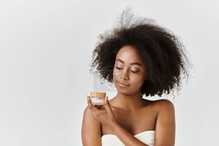 Une jeune afro-américaine aux cheveux bouclés se tient dans une serviette, tenant la crème en studio.