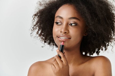 Eine schöne junge Afroamerikanerin mit lockigem Haar hält einen Lippenstift in der Hand und konzentriert sich auf Schönheit.