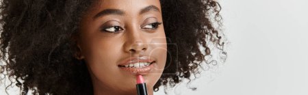 Una hermosa y joven mujer afroamericana con el pelo rizado sostiene elegantemente un tubo de lápiz labial en un entorno de estudio