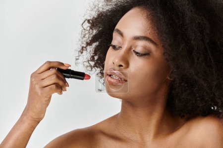 Foto de Una joven afroamericana con el pelo rizado se está aplicando hábilmente lápiz labial en la cara en un ambiente de estudio. - Imagen libre de derechos