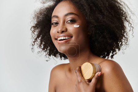 Une belle jeune femme afro-américaine aux cheveux bouclés tient une crème avec couverture en bois dans un cadre studio, exsudant un concept de soins de la peau.