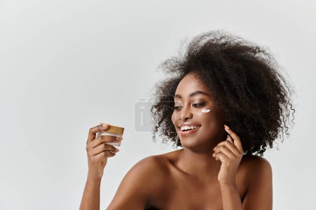 Téléchargez les photos : Une jeune afro-américaine aux cheveux bouclés sourit tout en tenant un pot de crème, rayonnant de joie et de contentement. - en image libre de droit