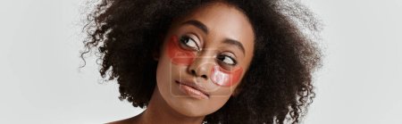 A stunning young African American woman with curly hair embodies a skin care concept while sporting a striking red eye patch.