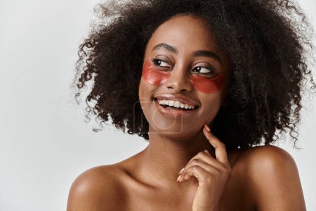 Foto de Una joven y feliz mujer afroamericana con el pelo rizado usando un llamativo parche rojo en un ambiente de estudio. - Imagen libre de derechos