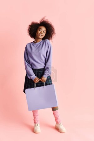 Foto de Una hermosa joven afroamericana con el pelo rizado sonriendo mientras sostiene una bolsa de compras en un entorno de estudio. - Imagen libre de derechos
