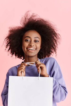 A beautiful young African American woman with curly hair happily holds a shopping bag in a studio setting.