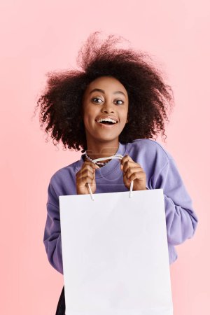Foto de Una hermosa joven afroamericana con el pelo rizado sosteniendo una bolsa de compras y sonriendo en un ambiente de estudio. - Imagen libre de derechos