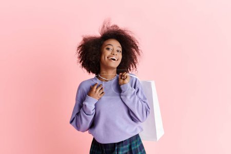 Photo for A young African American woman with curly hair holding a shopping bag and smiling in a studio setting. - Royalty Free Image