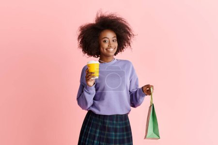 Photo for A stylish African American woman with curly hair holds a cup of coffee and a paper bag in a fashionable studio setting. - Royalty Free Image