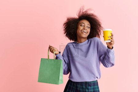 Foto de Hermosa mujer afroamericana con el pelo rizado sosteniendo una taza de café y una bolsa de papel, disfrutando de una juerga de compras por la mañana. - Imagen libre de derechos