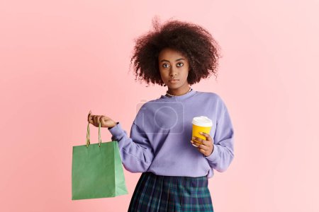 Foto de Una mujer afroamericana elegante con el pelo rizado sostiene una taza de café y una bolsa de papel. - Imagen libre de derechos