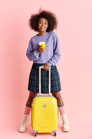 A beautiful young African American woman with curly hair holds a yellow suitcase and a cup of coffee, ready for travel.