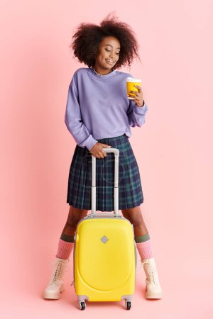 Young African American woman in purple sweater and plaid skirt, holding yellow suitcase in studio setting.