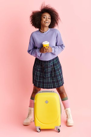 A young African American woman stands with a suitcase, holding a cup of coffee in a stylish studio setting.
