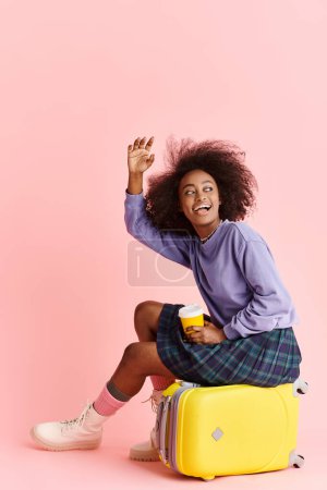 Foto de A beautiful young African American woman sits atop a bright yellow suitcase in a studio setting, exuding a sense of wanderlust and fashion. - Imagen libre de derechos