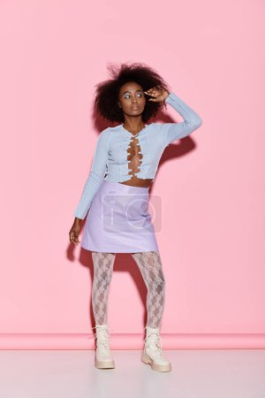 Foto de Una joven y elegante mujer afroamericana con el pelo rizado está de pie con confianza frente a una pared de color rosa brillante en un entorno de estudio. - Imagen libre de derechos