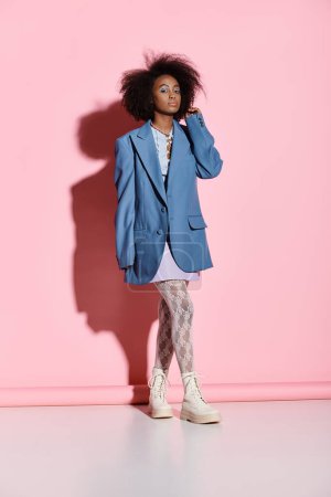 Téléchargez les photos : Une jeune femme afro-américaine élégante aux cheveux bouclés pose devant un mur rose vif dans un décor de studio. - en image libre de droit