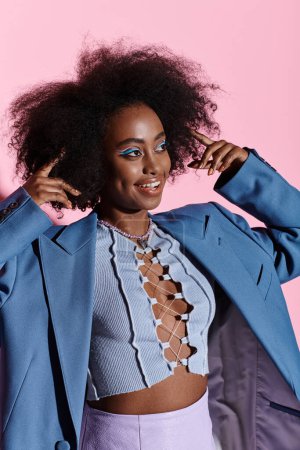 A stylish young African American woman with curly hair poses in a studio wearing a striking blue jacket for a photograph.